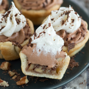 **Mini Bocconcini di Torta alle Noci Pecan e Cioccolato Vellutato** 5 Mini tortine noci pecan e cioccolato silk guarnite con spirali di cioccolato e panna montata, decorate con scaglie di cioccolato, disposte su un piatto. Una tortina è tagliata a metà, rivelando un ripieno di noci all'interno della crosta friabile.