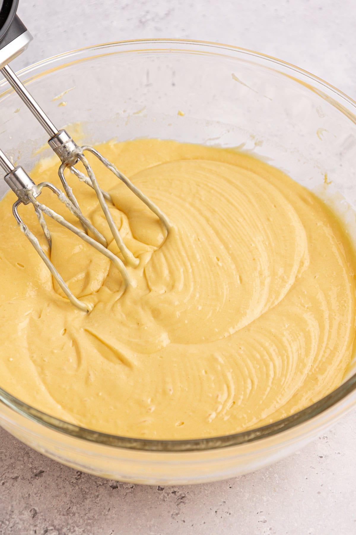 Torta Girella al Miele 6 Una ciotola di vetro piena di impasto giallo liscio e cremoso, mescolato da un frullino elettrico a mano con due fruste. La superficie dell'impasto della torta Honey Bun è texturizzata con spirali dovute al processo di miscelazione. Lo sfondo è una superficie chiara e maculata.