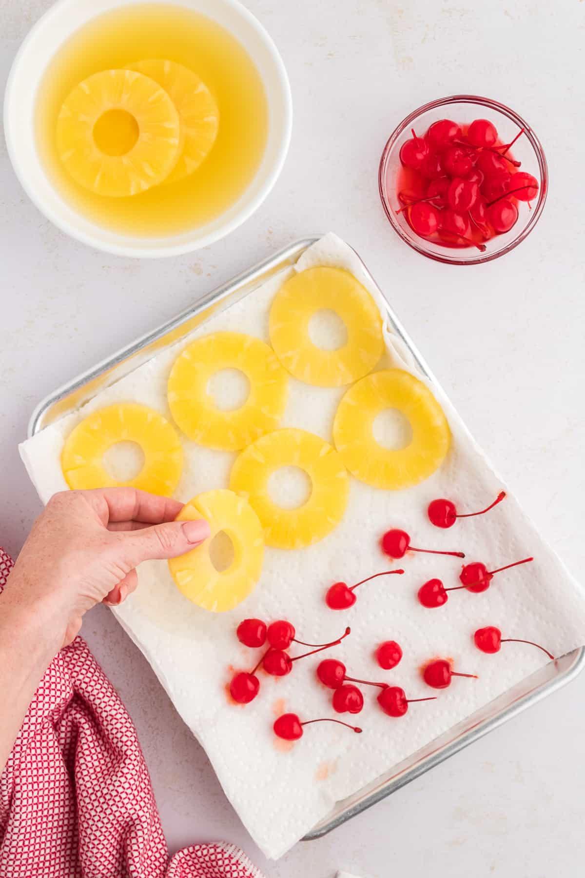 Torta di Ananas Rovesciata Fatta in Casa 6 Una teglia rivestita di carta assorbente con fette di ananas e ciliegie al maraschino, accanto a un canovaccio rosso e bianco, con una mano che aggiunge un'altra fetta di ananas, accanto a una ciotola di ciliegie e una ciotola di ananas.