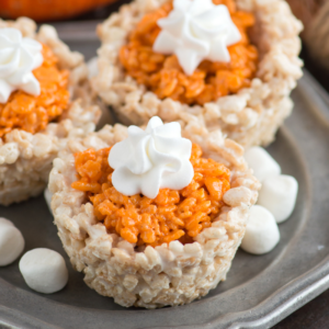**Mini Dolcetti di Riso Soffiato Gusto Torta di Zucca** 6 Tre dolcetti Rice Krispies alla zucca a forma di tazza, riempiti con una miscela di marshmallow arancione e guarniti con un vortice di panna montata bianca, disposti su un vassoio di metallo con mini marshmallows sparsi intorno.