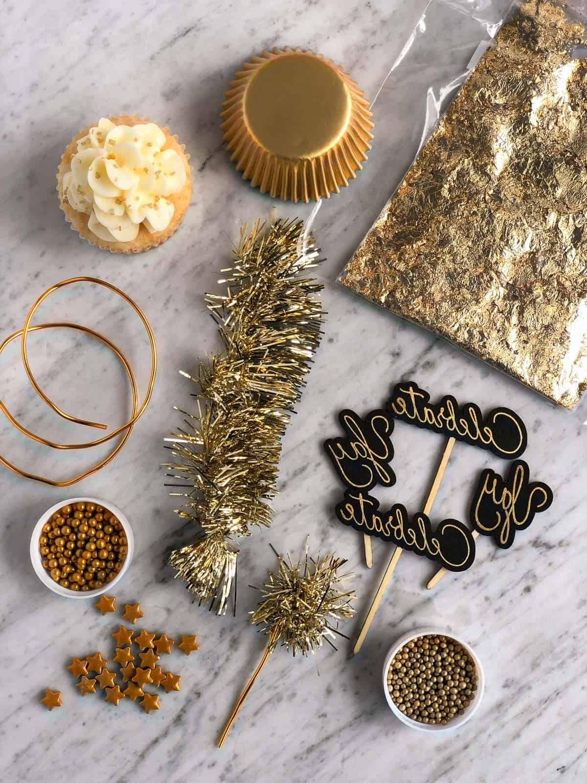 Questa ricetta facile per cupcakes allo champagne con glassa allo champagne è un dessert di Capodanno da non perdere! Come fare i cupcakes allo champagne usando una torta in scatola. Forniture utilizzate per decorare i cupcakes di Capodanno, tra cui zuccherini dorati, stelle dorate, pirottini dorati, foglia d'oro commestibile, topper per cupcake con la scritta 'celebrate' e tinsel dorato