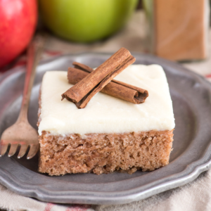 Torta Pazza alla Cannella e Spezie 2 Una fetta quadrata di crazy cake con glassa bianca, guarnita con due stecche di cannella, su un piatto grigio accanto a una forchetta. Mele e una stecca di cannella sono sfocate sullo sfondo.