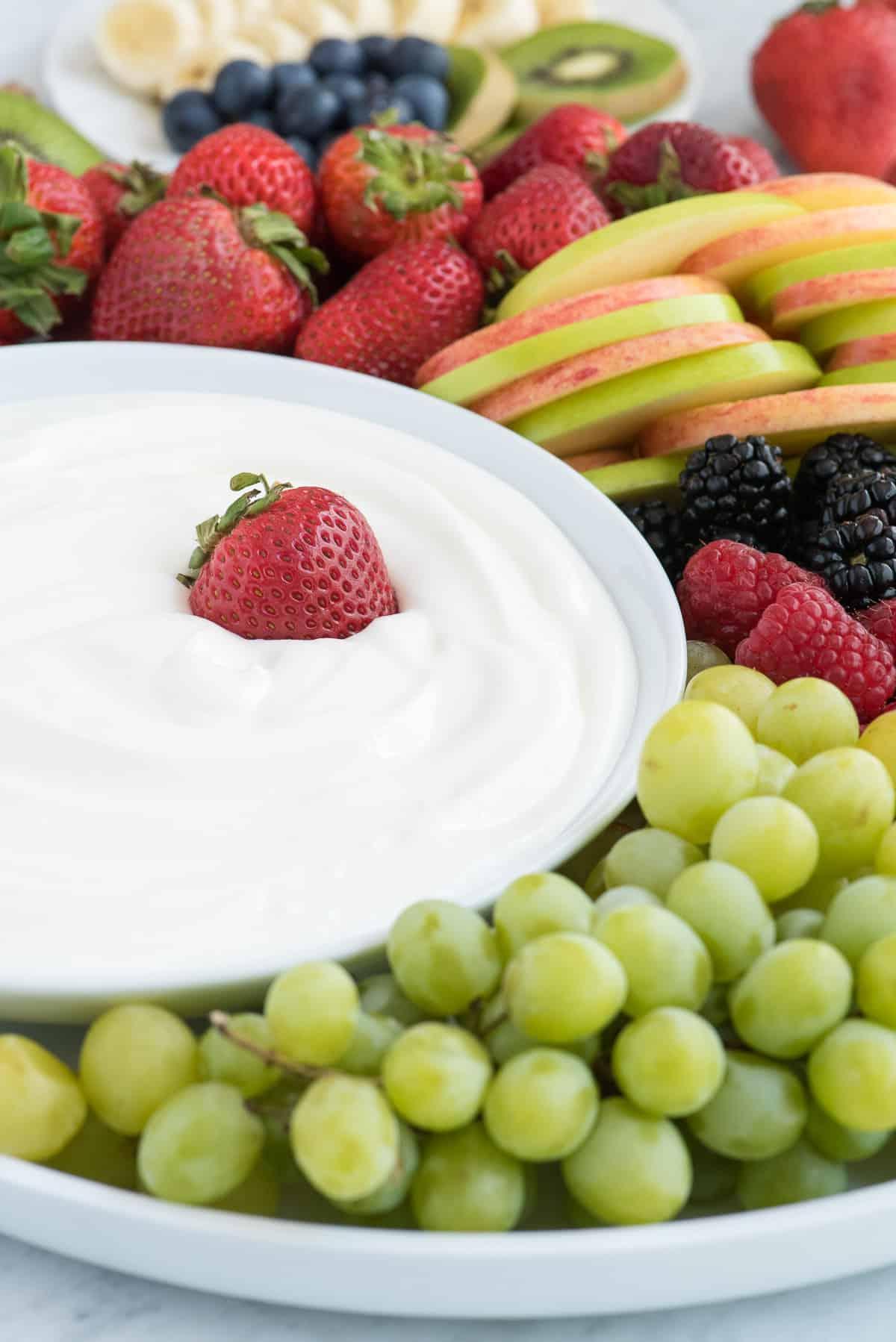 Dip alla frutta con 3 ingredienti! Amiamo questa crema vellutata perché NON c'è formaggio spalmabile! Piatto rotondo pieno di fragole, mele, more, lamponi e uva con una ciotola di dip bianco per frutta al centro