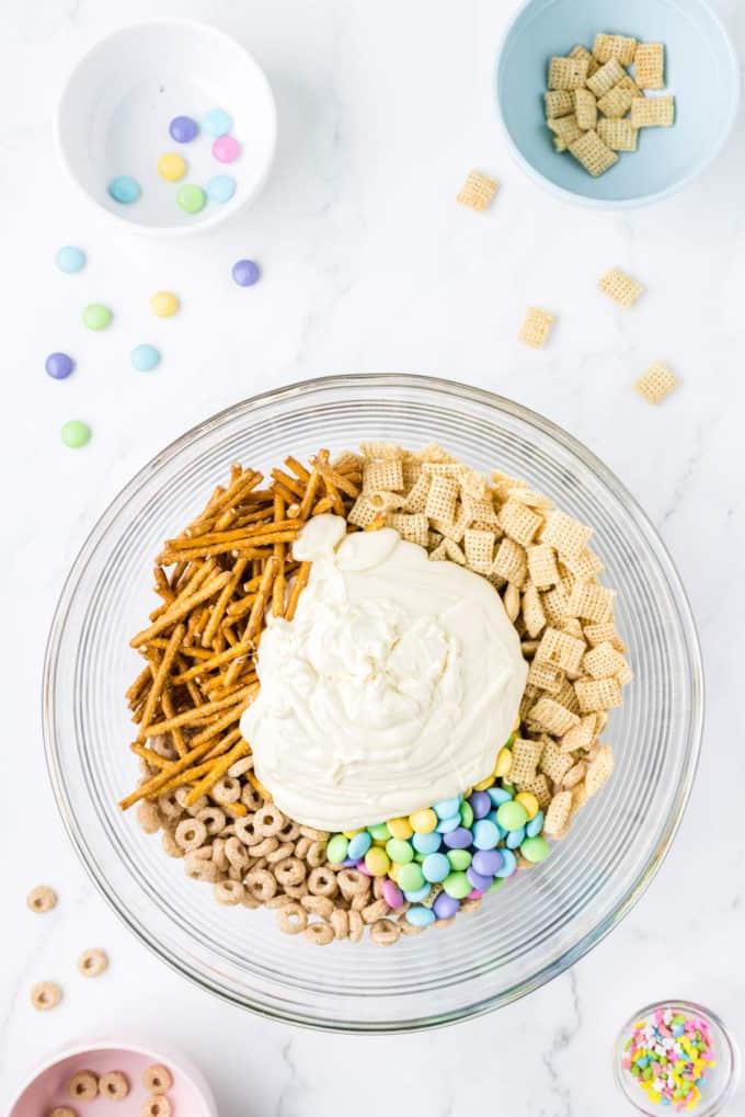 Esca per conigli 6 Ingredienti per 'Bunny Bait' in una ciotola di vetro trasparente circondata da una ciotola di codette, una ciotola con Cheerios, una ciotola di Chex di riso e una ciotola di M&M's