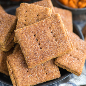 Biscotti Graham alla Zucca 7 Primo piano di un piatto pieno di biscotti Graham alla zucca rettangolari, cosparsi di cannella e zucchero, ognuno con piccoli fori al centro. Alcuni biscotti sono impilati, mostrando la loro consistenza croccante e dorata.