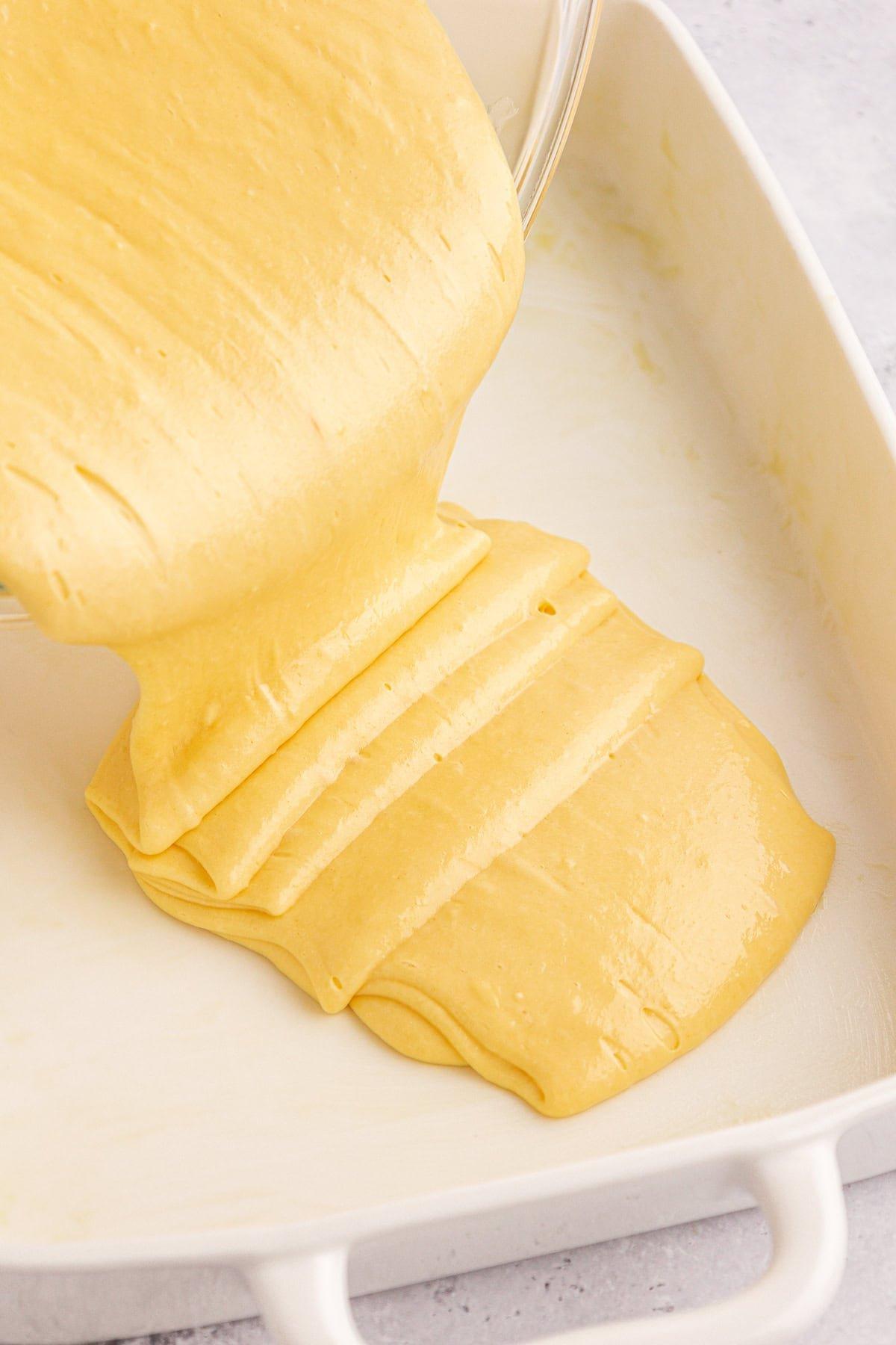 Torta Girella al Miele 7 L'impasto giallo della torta viene versato da una ciotola in una teglia rettangolare bianca, destinata a trasformarsi in una deliziosa torta Honey Bun. L'impasto liscio e lucido si stende uniformemente sul fondo del piatto, promettendo una dolce perfezione.