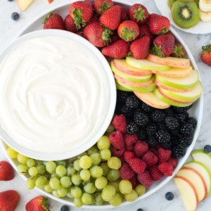 Crema di Frutta con Panna Montata 2 Un vassoio con fragole fresche, fette di mela, more, lamponi, uva verde e una ciotola di cremoso dip alla frutta con yogurt al centro, esposto su una superficie di marmo.