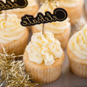 Cupcakes Facili allo Champagne 3 Champagne cupcakes con spirali di glassa bianca, decorati con zuccherini dorati e un cartello a stuzzicadenti con la scritta “Celebrate”. Decorazioni in tinsel dorato sono posizionate intorno ai cupcakes.