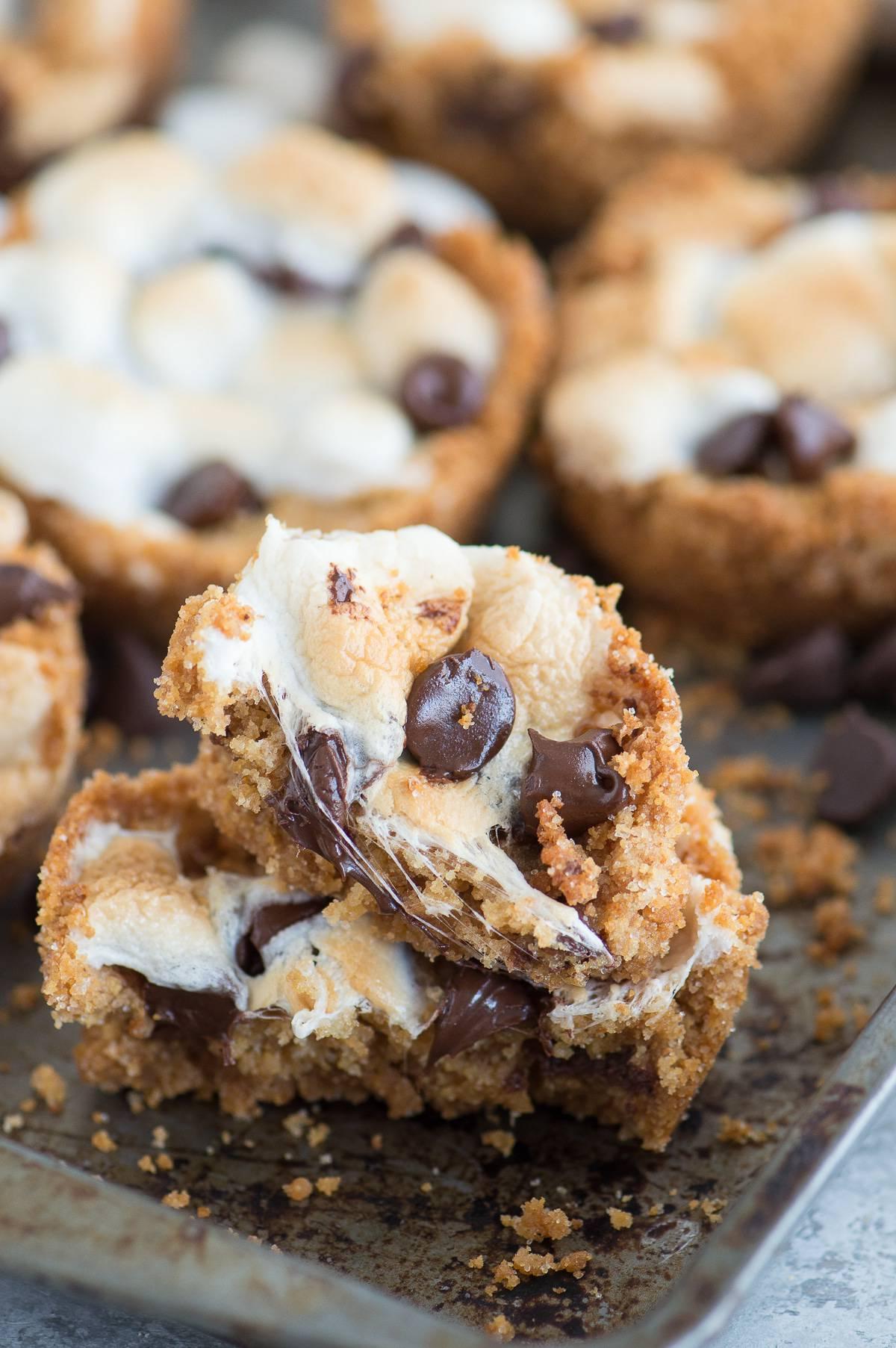 Tazze S'mores al Forno con 5 ingredienti! Preparale in uno stampo per muffin standard. Un incrocio tra biscotti s'mores e cupcake s'mores! Due Tazze S'mores da Forno mezze mangiate circondate da altre tazze s'mores fatte in casa su una teglia da forno.