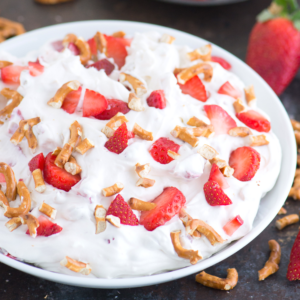 Insalata Soffice di Fragole e Salatini 7 Una ciotola di dessert cremoso guarnito con fragole a fette e pezzi di pretzel rotti, con una fragola intera e altri pretzel sparsi nelle vicinanze su una superficie scura, pronto per essere gustato.