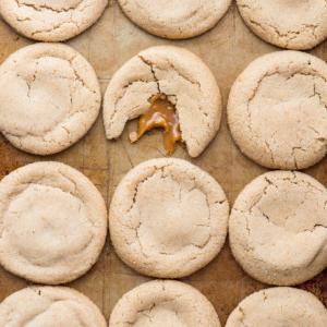 Biscotti alle Mele e al Caramello 8 Twelve round caramel apple cookies on a baking sheet; one cookie in the center is broken, revealing gooey caramel inside.