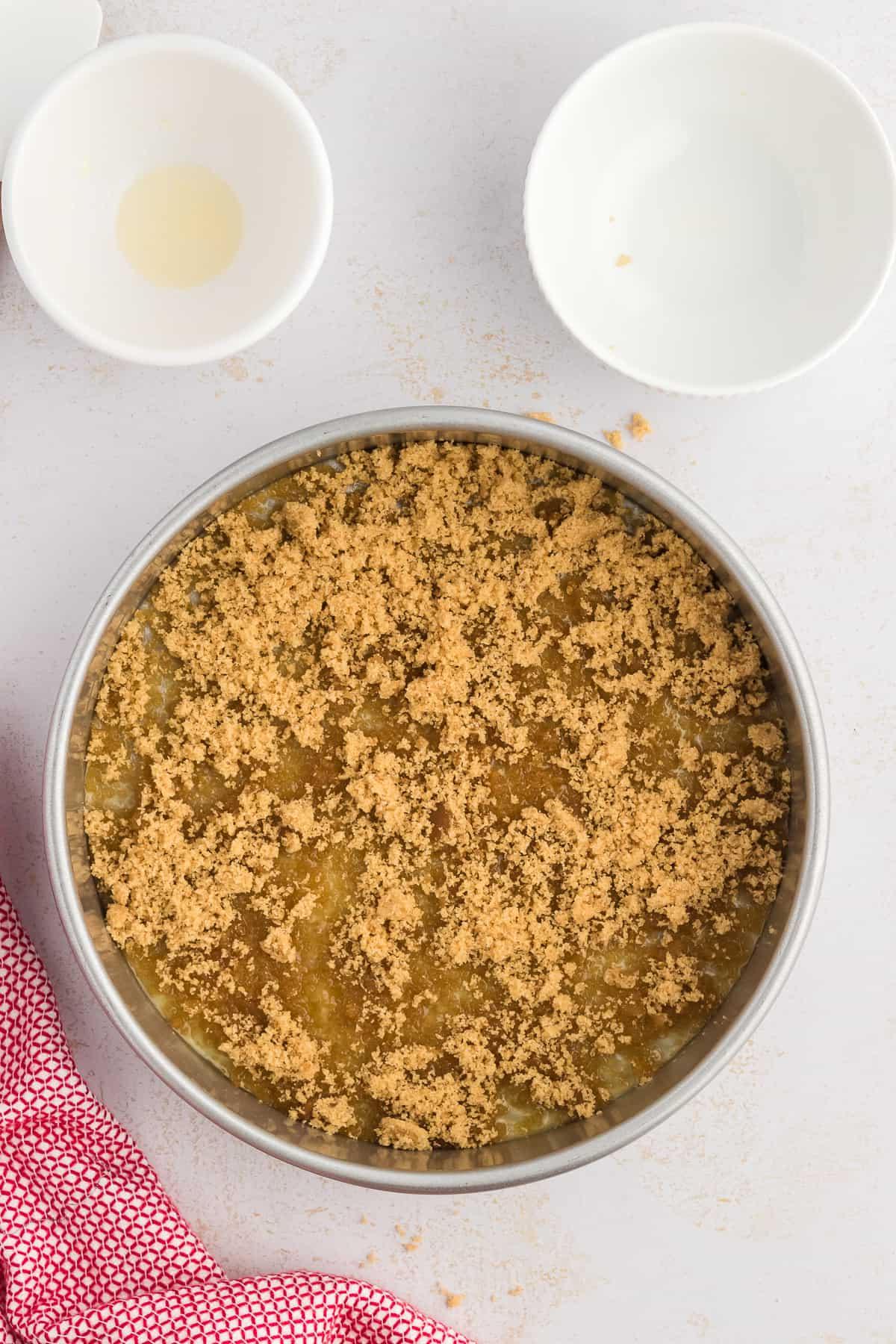 Torta di Ananas Rovesciata Fatta in Casa 8 Una teglia rotonda per torte con burro fuso e zucchero di canna nel fondo, circondata da due piccole ciotole bianche vuote e un canovaccio rosso e bianco.