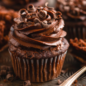 Cupcakes al cioccolato 8 Primo piano di ricchi cupcake al cioccolato guarniti con glassa al cioccolato a spirale e scaglie di cioccolato, con un altro cupcake sfocato sullo sfondo.
