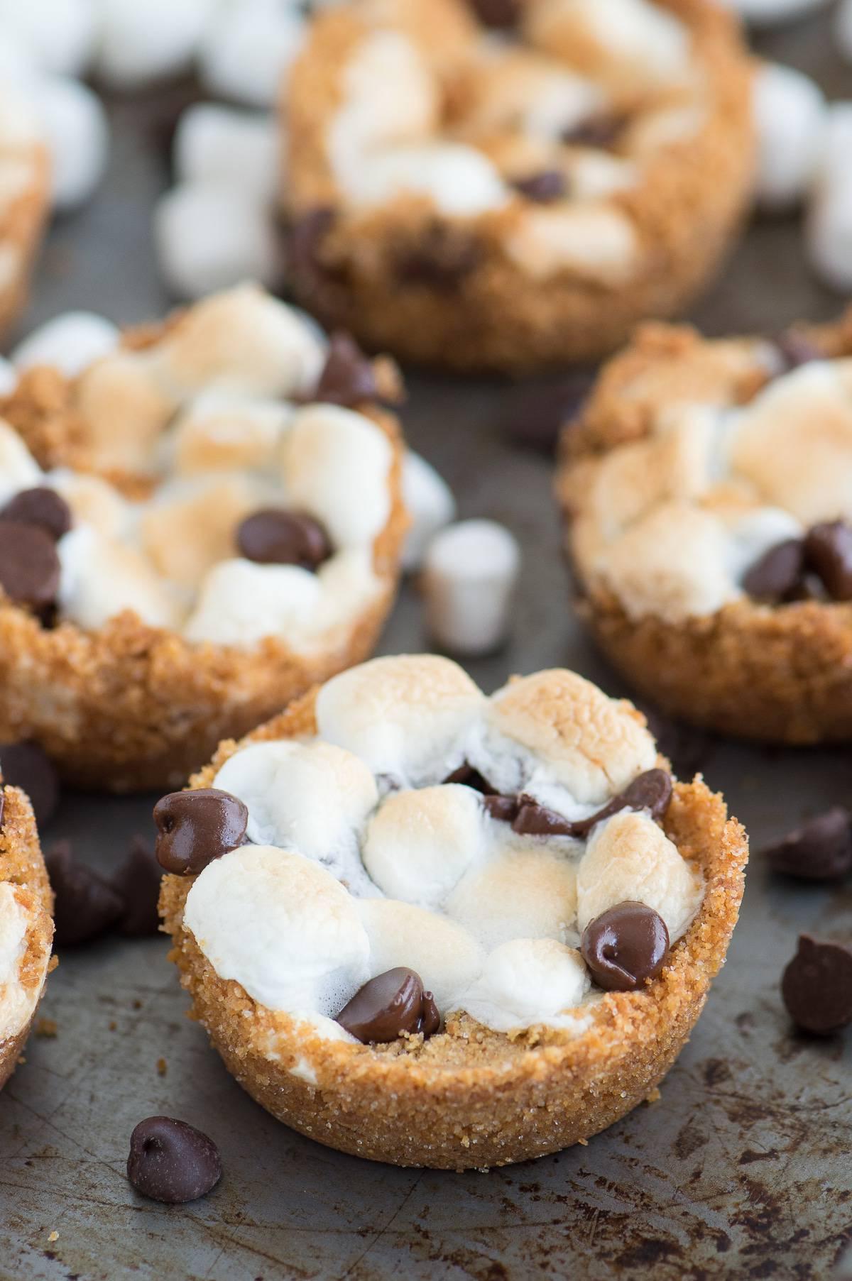 Tazze S'mores al Forno con 5 ingredienti! Preparale in uno stampo per muffin standard. Un incrocio tra biscotti s'mores e cupcake s'mores! Disposizione di quattro Tazze S'mores da Forno con 5 ingredienti su una teglia.