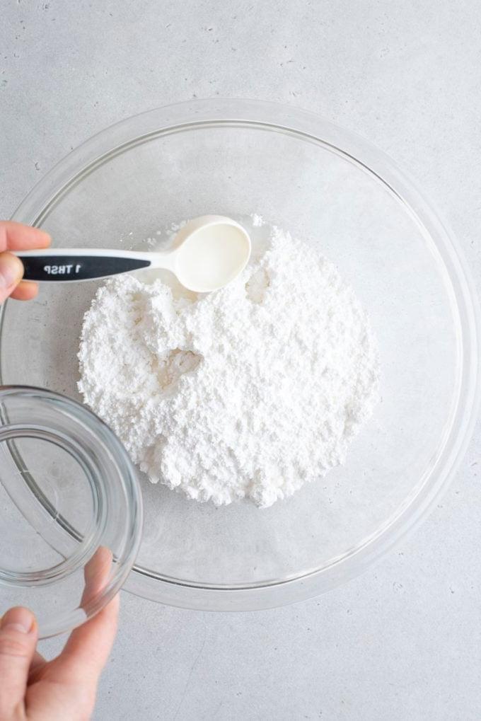 Glassa Reale senza Polvere di Meringa 8 ingredienti per ghiaccia reale in una ciotola di vetro trasparente con acqua aggiunta usando un piccolo cucchiaino dosatore