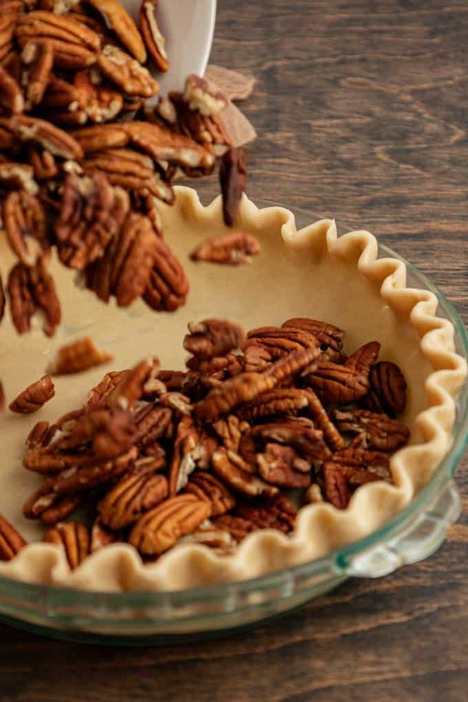 Torta di Noci Pecan Casereccia All'Antica 8 Noci pecan aggiunte a una teglia di vetro con crosta di torta non cotta, poggiata su una superficie di legno