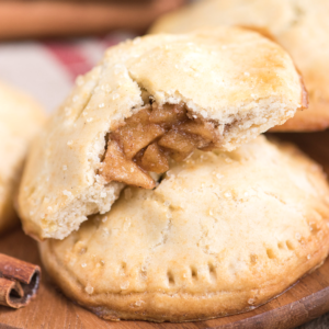 Fagottini di mele fatti in casa 2 Una tortina di mele dorata e croccante in primo piano, rotta per rivelare fette di mela speziate. La mini torta poggia su una superficie di legno con bastoncini di cannella sullo sfondo.