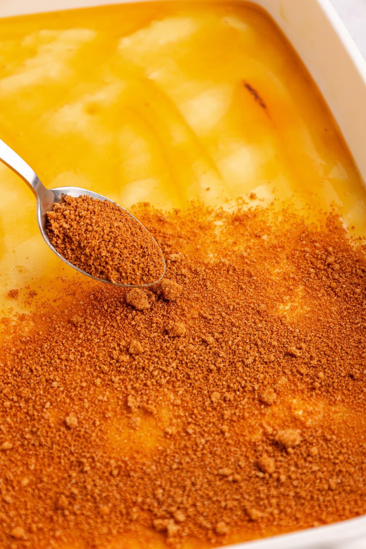 Torta Girella al Miele 9 Un cucchiaio sparge zucchero di canna su una torta Honey Bun gialla cremosa in una teglia rettangolare. Il dessert sembra avere una consistenza liscia, contrastante con la guarnizione granulata.