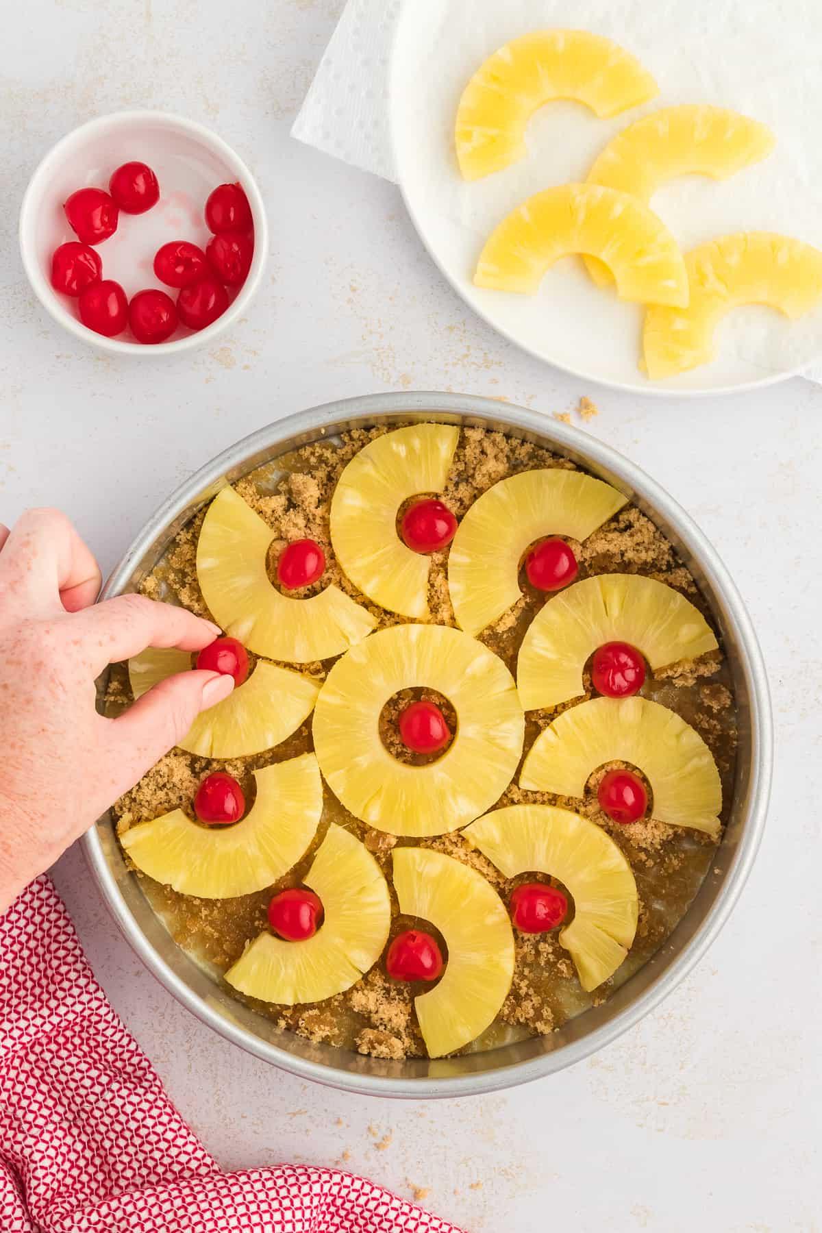 Torta di Ananas Rovesciata Fatta in Casa 9 Una teglia per torte con zucchero di canna e burro fuso nel fondo e una mano che sistema fette di ananas e ciliegie al maraschino, accanto a un canovaccio rosso e bianco, un piatto di ananas a fette e una piccola ciotola di ciliegie.