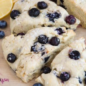 I Migliori Scones al Limone e Mirtilli 10 Tre scones triangolari al limone e mirtilli con una leggera spolverata di zucchero sono disposti su carta forno, circondati da mirtilli freschi e fette di limone sullo sfondo.