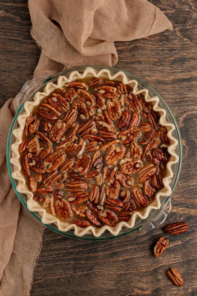 Torta di Noci Pecan Casereccia All'Antica 10 Una torta di noci pecan pronta per essere cotta in una teglia di vetro, poggiata su una superficie di legno con un telo color tortora e noci pecan intere intorno