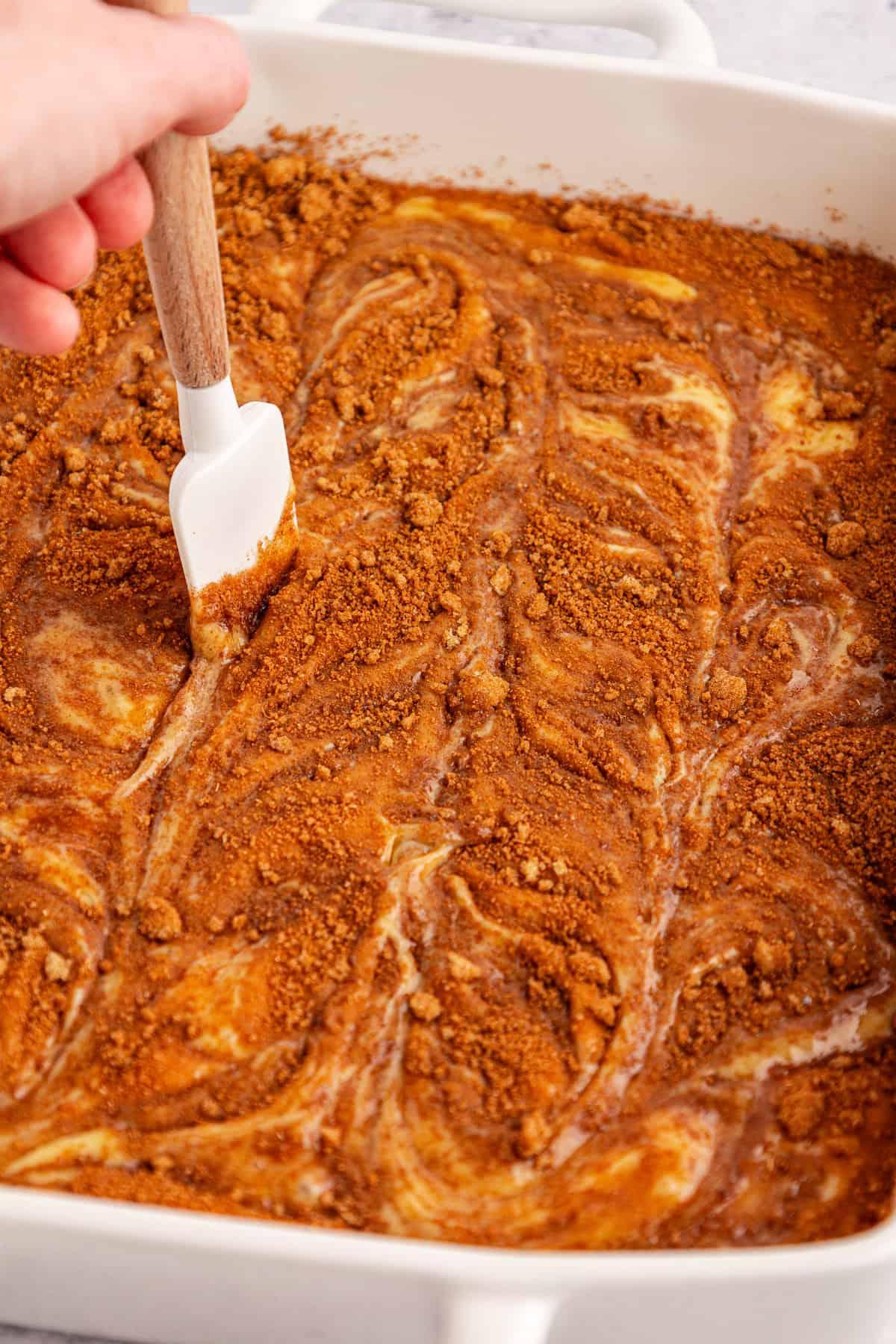 Torta Girella al Miele 10 Una mano muove delicatamente una spatola bianca nell'impasto della torta Honey Bun, composto da cannella, zucchero di canna e impasto, in una teglia da forno bianca, creando un bellissimo effetto marmorizzato.