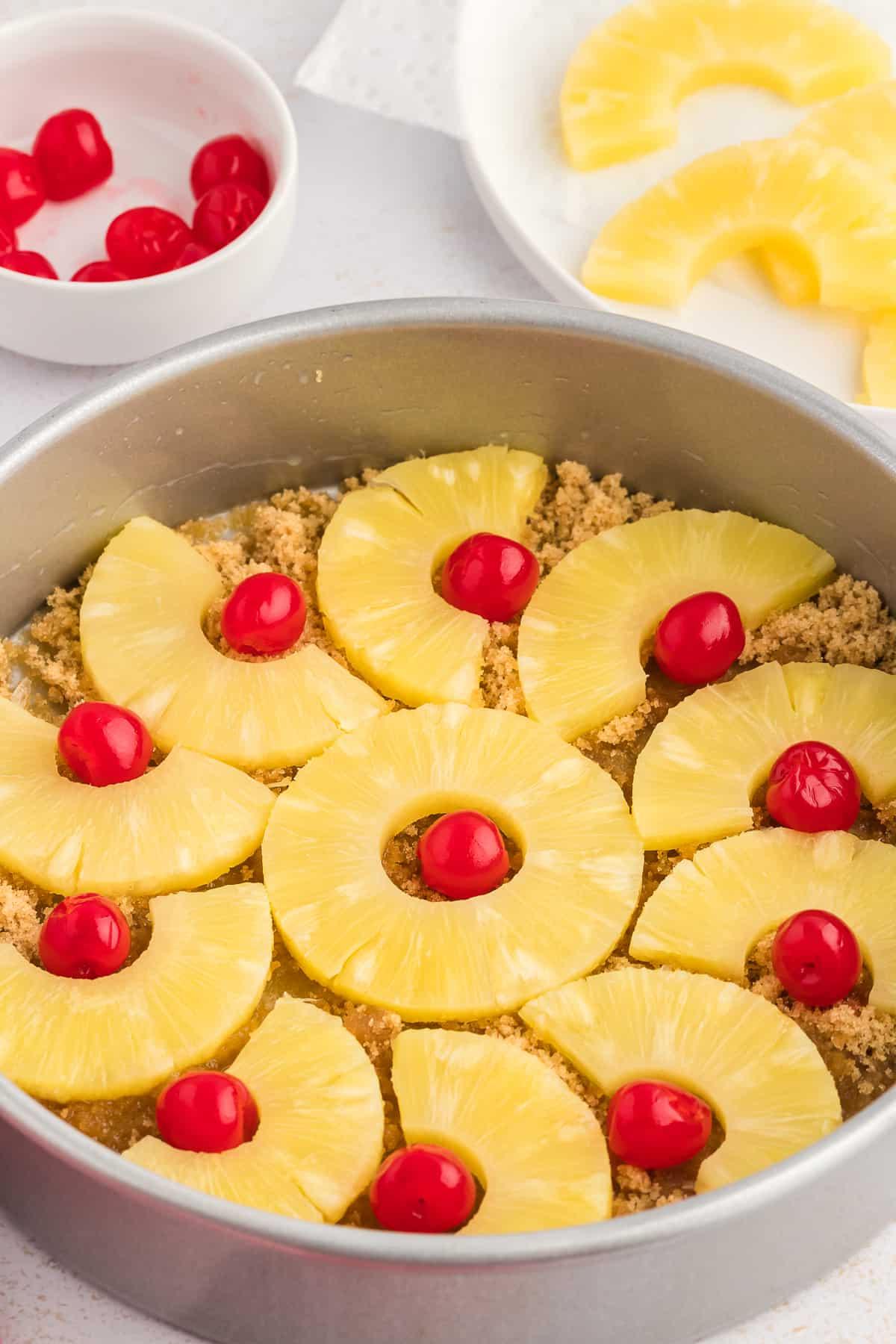 Torta di Ananas Rovesciata Fatta in Casa 10 Una teglia per torte con zucchero di canna e burro fuso nel fondo e fette di ananas e ciliegie al maraschino disposte, accanto a un canovaccio rosso e bianco, un piatto di ananas a fette e una piccola ciotola di ciliegie.