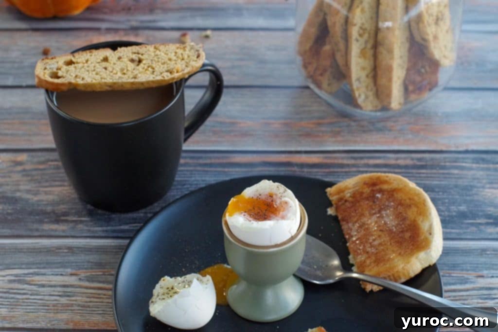 Pumpkin pecan biscotti resting on a cup of coffee, with a plate of breakfast items like a boiled egg and toast in the foreground.