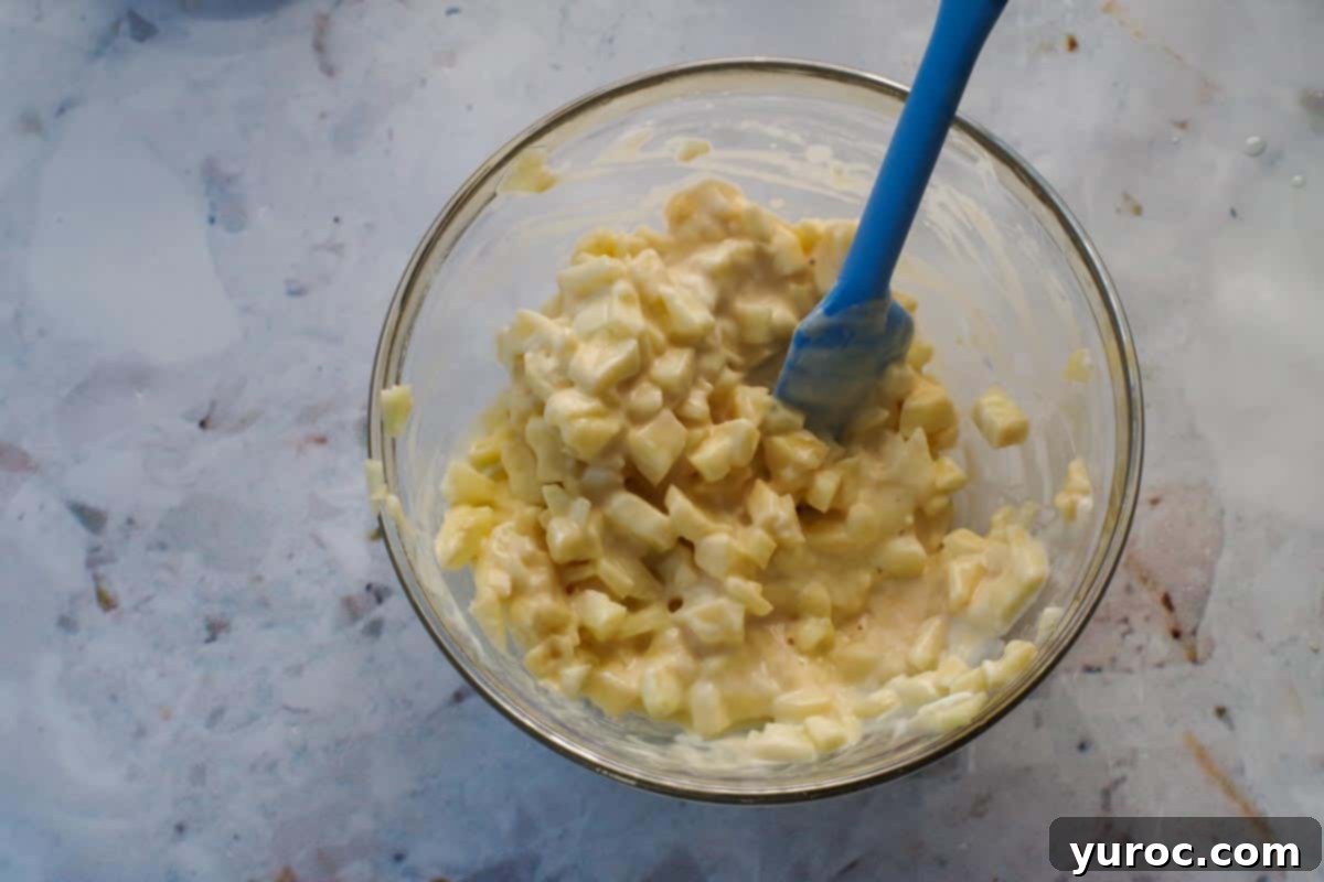 Yogurt and vanilla mixed into egg mixture with apple chunks in medium glass bowl with blue spatula.