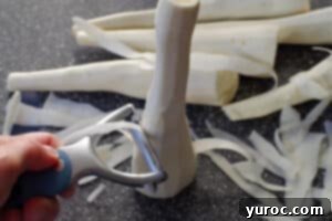 Parsnip being ribboned with a vegetable peeler on a cutting board, with additional parsnips in the background.