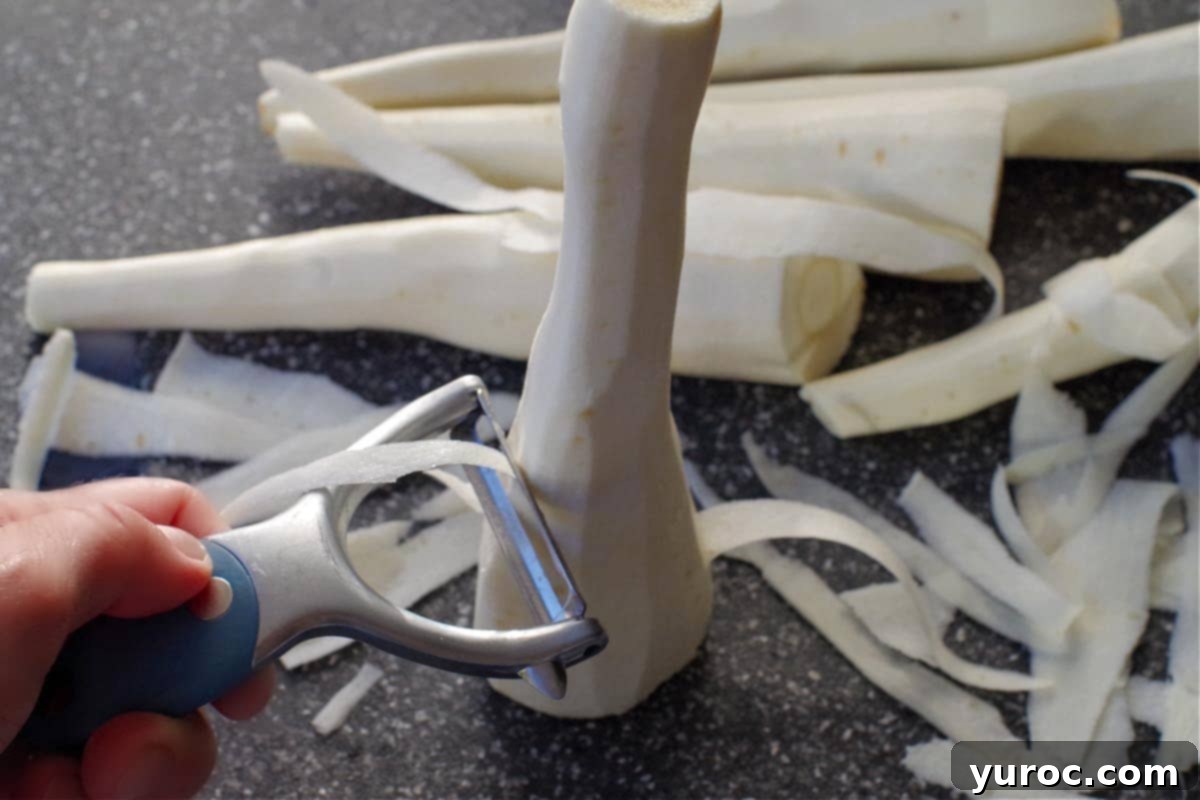 Hands demonstrating how to ribbon a parsnip with a vegetable peeler on a wooden cutting board, with more parsnips in the background ready for preparation.