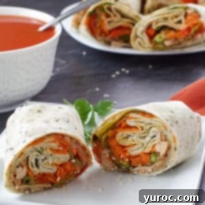 healthy turkey wrap, sliced in half, on white plate, with bowl of tomato soup and platter of turkey wraps in the background