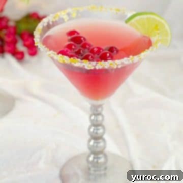 Cranberry Cosmopolitan Mocktail with cranberries floating in drink, wedge of lime and sparkly gold sugar with cranberries in the background, on white material