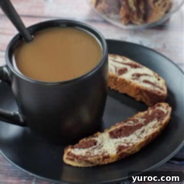 cup of coffee in a black mug on a black plate with 2 pieces of chocolate marble biscotti on the plate and a jar of biscotti in background