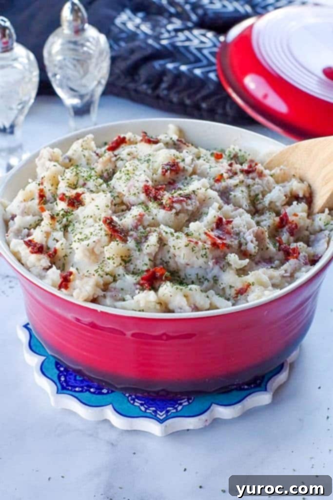 Chipotle mashed potatoes in a red serving bowl with a wooden spoon, with black oven mitts and crystal salt and pepper shakers in the background.