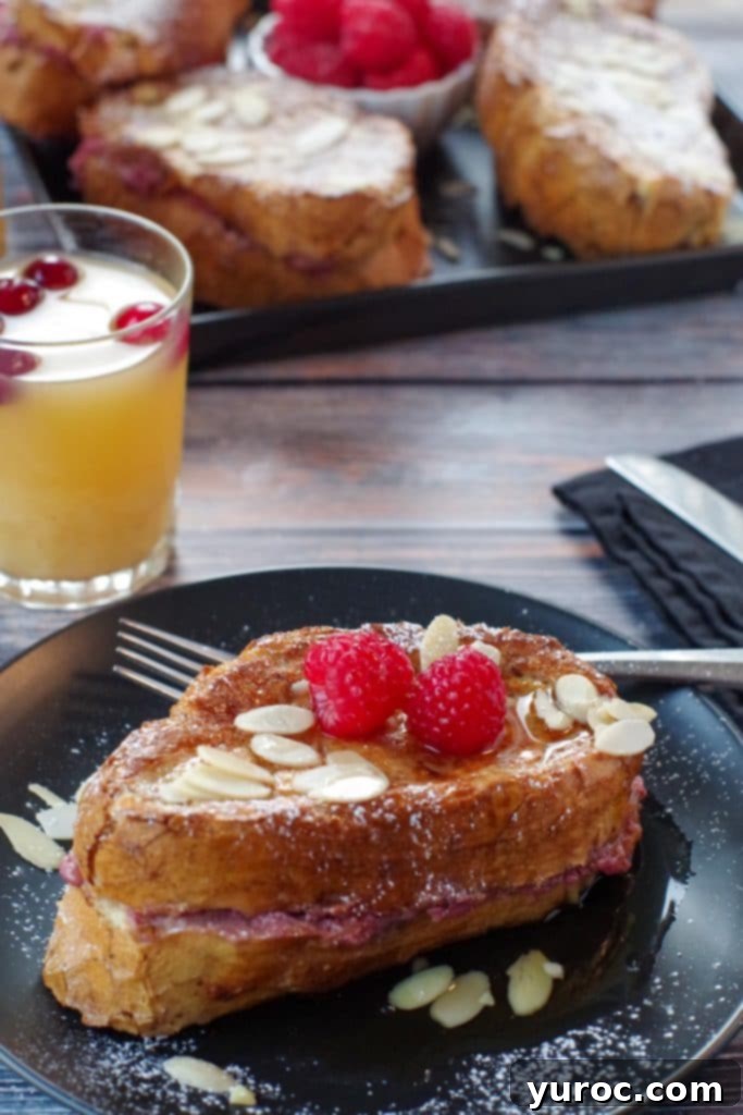 Raspberry Cream-Filled French Toast Sandwich 2 French Toast Sandwich on a black plate with fork and glass of juice and tray of more sandwiches in background