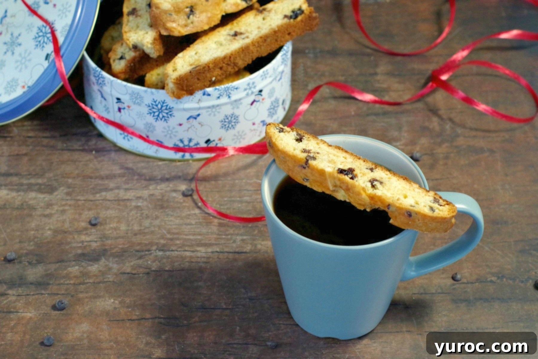 Black Forest biscotti cookies arranged on a blue mug with a festive Christmas tin of more biscotti in the background, creating a cozy holiday scene.