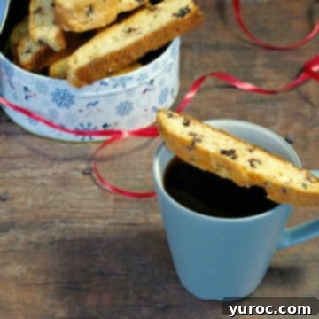 Decadent Black Forest Biscotti 8 Black Forest Biscotti cookies, perfect for dipping in coffee, arranged on a blue mug with a festive tin of more biscotti in the background.