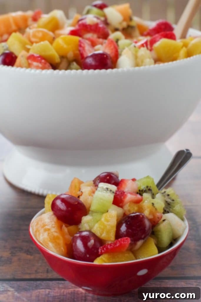 Classic fruit salad presented in a small red bowl with white polka dots, with a large white bowl of the same salad in the soft-focus background.