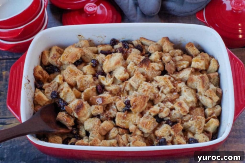 Festive Cranberry Bread Pudding 3 bread pudding with condensed milk in a red casserole dish the a wooden spoon in it and red dishes and grey oven mitts in background