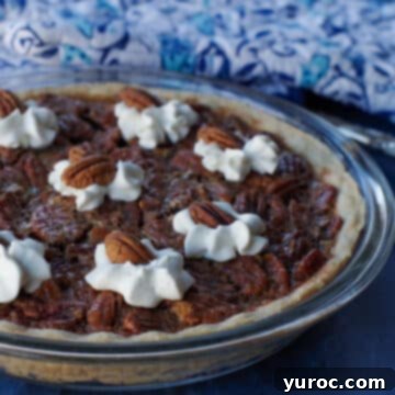 A golden-brown pecan pie in a glass pie dish, resting on a rustic blue surface, with blue patterned oven mitts casually placed in the background.