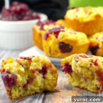 A perfectly split cranberry cornbread muffin revealing its tender interior, with a plate of more delicious muffins in the background.