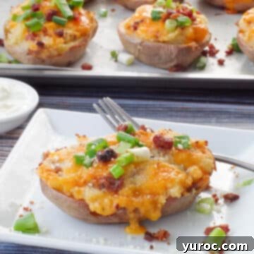 A perfectly baked Caesar twice baked potato resting on a white plate with a fork and a small dish of sour cream, with a sheet pan of more potatoes in the background.