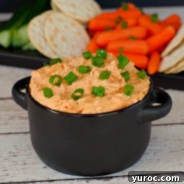 Creamy buffalo chicken dip served in a black container, with a vibrant array of fresh vegetables and crackers artfully arranged in the background.