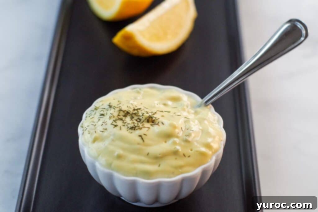 Freshly made healthy tartar sauce in a small white dish with a spoon, accompanied by sliced lemons on a dark tray, highlighting its vibrant, inviting appearance.