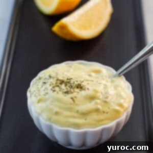 Freshly made healthy tartar sauce in a small white dish with a spoon, accompanied by sliced lemons on a dark tray.