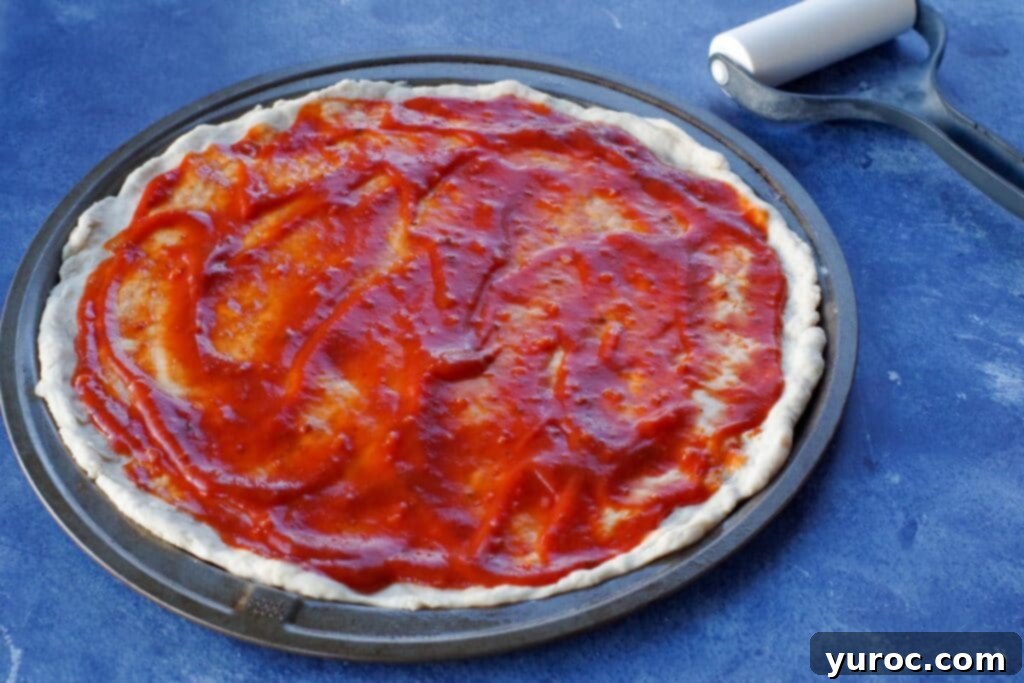 Şipşak 15 Dakika Pizza Hamuru 2 pizza dough with sauce on pan, on blue counter top surface with rolling pin in background