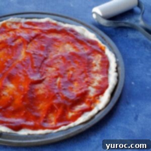 Şipşak 15 Dakika Pizza Hamuru 10 pizza dough with sauce on pan, on blue counter top surface with rolling pin in background