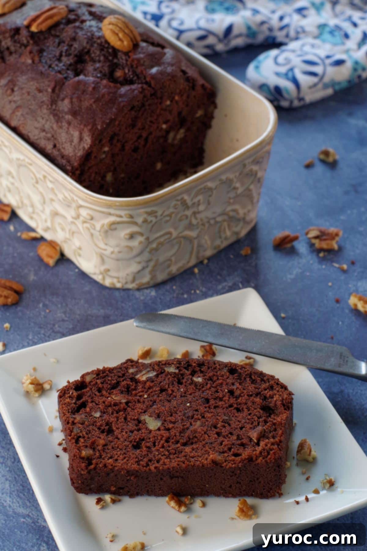 Blender Bliss Chocolate Zucchini Bread 1 A slice of moist chocolate blender zucchini bread on a white plate with a knife, and the rest of the loaf in the background, showcasing its rich, dark color and tender crumb.