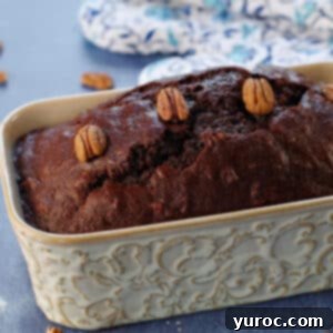 Blender Bliss Chocolate Zucchini Bread 16 An entire, perfectly baked loaf of chocolate zucchini bread, with blue patterned oven mitts in the background and a scattering of pecans around, indicating fresh out of the oven goodness.